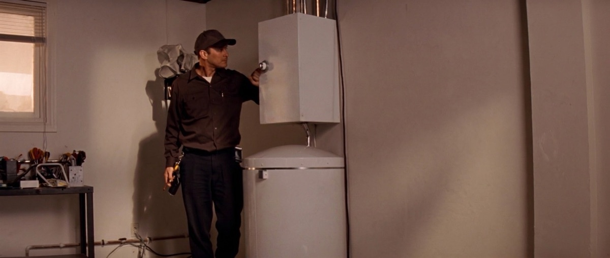An HVAC technician in a dark uniform adjusts the control valve on a residential water heater in a home utility room